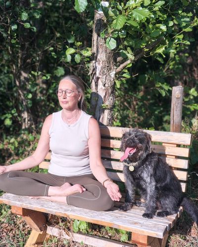 Heike meditiert auf einer Holzbank am Waldrand, neben ihr ein Hund